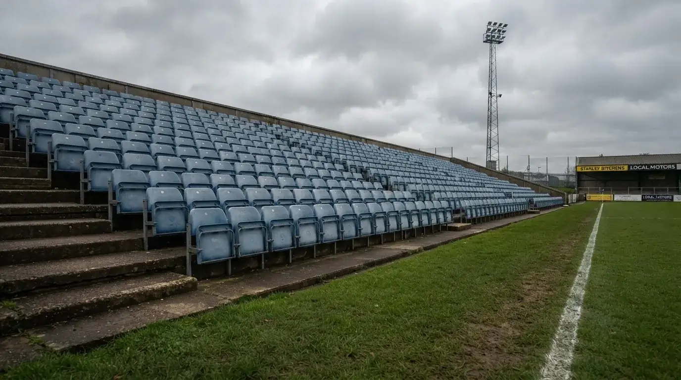 Rzędy pustych plastikowych krzesełek małego stadionu widziane z poziomu murawy pod pochmurnym niebem.